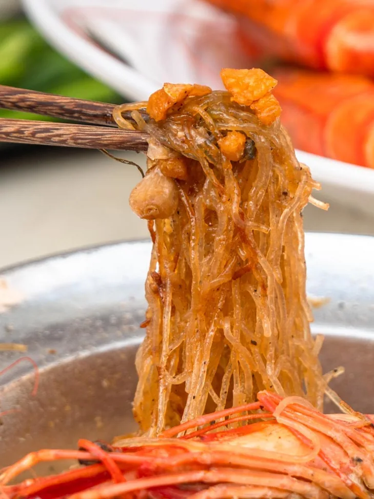 Chopsticks lifting glass noodles from a steelpot with king prawns